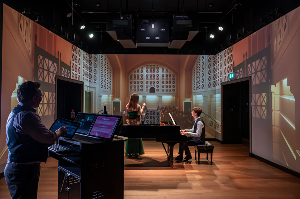 Two students, one student singing and one student playing the piano, performing in front of a fake audience in a simulator, with a man managing a tablet and screens.